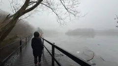 Weather Monitor/Raj's Range A man wearing a dark parka-style coat is standing on a wooden bridge. They see an icy lake in the distance amidst the fog and trees are visible