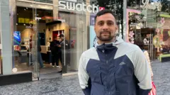 A man, in his late 20s or early 30s, with black hair and a black beard, smiles at the camera outside a glass-fronted shop with the Swatch logo above the door in illuminated white lettering. He is wearing a grey and blue rain coat. 