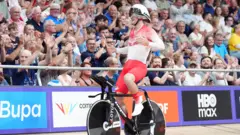 A picture of Will Tidball smiling. He is wearing a white cycle helmet and a Team England red and white racing outfit while astride a black bike. He next to a large crowd in a velodrome.