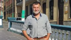Michael Bier A man with grey beard and short greying brown hair is standing in a town square. He is a wearing a faded, blue and white striped shirt and light brown chinos with dark brown belt. He has a relaxed pose. 
