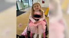 Charlie Brown Danny-Ray Brown is wearing a tiara and a pink princess dress, and holding a cane, while she sits in a wheelchair in a room inside the house.
