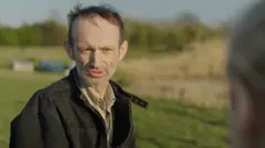 Andi Miller, a man with thin, brown hair and greying stubble, wearing a dark jacket and a collared shirt and sitting in a rural location with trees and fields behind him as he is being interviewed.