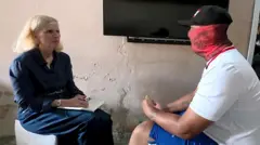 Orla Guerin speaks to a man wearing a black baseball cap and red scarf covering his face.