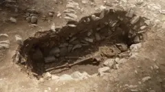 Kevin Church/BBC News A human skeleton is lying in an oval-shaped grave cut into limestone bedrock. The bones are positioned naturally, with the skull at one end and legs extended. The ground surrounding the grave is light brown and cracked.