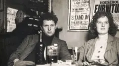 Getty Images Dylan Thomas sits with his wife, Caitlin, at a pub table with a stack of beers. He is wearing a checkered jacket and is looking straight at the camera. Caitlin is looking to the side, her curly hair is open and she is wearing a jacket with a buttoned top underneath.