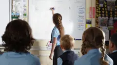 A primary school pupil in a blue and grey uniform, standing and writing at a whiteboard while pupils watch on.