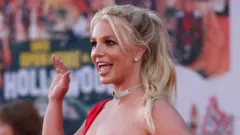 Reuters Britney Spears waves and smiles to a camera to the left, while at a premiere dressed in a red dress with a diamond choker necklace.
