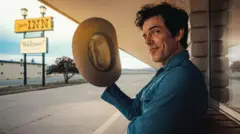 Brandon Flowers holds a stetson in his right hand and gives a wry look to the camera as he poses outside a stereotypical American motel