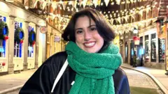 Samantha Rizzo Samantha smiles for the camera with a green scarf wrapped around her neck. He wears a black leather jacket and has brown hair. In the background we can see a festive street of shops decorated with flowers and garlands. It is evening time and the yellow fairy lights are on.