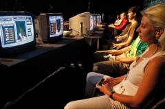 Cerca de 7 pessoas sentadas em frente a telas em ambiente escuro