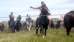 Native Americans on horseback ride down a mound in a field, towards the camera lens, re-enacting the battle of Little Bighorn