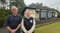 Barrie Bulch is wearing a blue polo shirt and is standing next to Councillor Paul Donaghy, who is wearing a black gilet with a blue Reform UK logo. They are standing on a bowling green in front of the burnt-out club house behind them.
