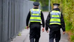 Two police officers are walking down a street. They are both wearing hi-vis vests over a black uniform and wearing black hats. There are bushes to their right and a metal fence to their left.