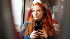 A women with red hair wearing headphones and holding her phone. She is sitting on a train and looking out of the window.