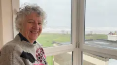 Helen Coyne is in her seventies and is wearing a beige jumper with a pattern of pink and purple flowers on it. She is standing by a window with white uPVC frames, through which some grass and the sea beyond can be seen. The day looks a bit grey.