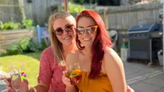 Leanne and Sarah/BBC Friends Sarah (left) and Leanne (right) wearing sunglasses, smiling and holding drinks as they sit in a garden.