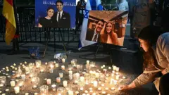 Dozens of lit candles shine on the sidewalk in front of two posterboards displaying photos of both the victims 