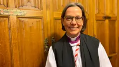 Bishop of Newcastle Helen Ann Hartley standing next to a wooden door. She wears her dog collar and a purple shirt, with a while and white robe. She had short dark bobbed hair and glasses.