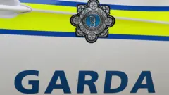 The side door of a Gardaí police car. The door is white with a yellow and blue luminous stripe across it. There is a grey and blue circle badge on the stripe. Below the stripe, in blue font, reads 'Garda'