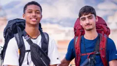 Jo and Kush, who are carrying large backpacks, are smiling at the camera at the start of Race Across The World in Italy. Jo is on the left and his wearing a white t-shirt with a black backpack, while Kush is wearing a blue t-shirt and has a red backpack. The background behind them is blurred but you can just about make out some blue sky and white clouds.