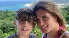 A mother and son pose for a selfie. They are both smiling with the tops of their heads touching. They are standing on a viewpoint, with a canopy of trees and an ocean view far below them.