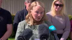 Geraldine Mullan is talking in front of mics. She's outside. She has shoulder length blond hair and is wearing a grey jacket. People are standing behind her.