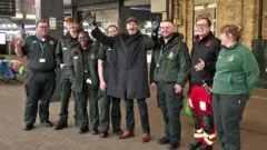 A beaming Ian Drewery stands with arms aloft in the middle of a group of eight men and women in uniforms on a station platform.