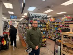 Carlos Macias stands in his small grocery store with his hands clasped in front of him. He wears an olive-green fleece sweatshirt