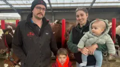 BBC Farmer Adam Johnstone, wearing a black gilet and black hat, and Lucy Johnstone, wearing a green fleece and black gilet, stand in front of a cowshed, with Lucy holding her 5-month-old son and her 4-year-old son standing between them.