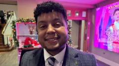 A mid shot of a man in a grey suit, white shirt and blue and striped tie. He has short dark curly hair and is standing in a foyer that is decorated with Christmas decorations. He is smiling.