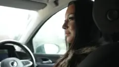 Alisa is sitting on the driver's seat of the car. She has long black hair and is wearing a furry coat