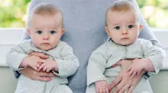 Two babies in green and white striped baby-grows. An adult is carrying them under either arm. 
