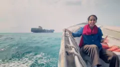 Nawal wears a scarf, shirt and trousers as she sits in a boat on the Strait with a shipping container seen in the background