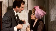 John Cleese and Prunella Scales as Basil and Sybil Fawlty in the 1970s. This is a scene from Fawlty Towers. Prunella Scales is wearing curlers and a hair cloth and she is arguing with John Cleese.