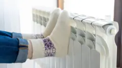 What's happening with gas and electricity prices? 15 Getty Images A woman wearing rolled-up blue jeans and white and purple knitted socks rests her feet on a white radiator.