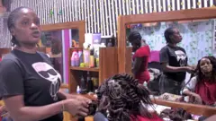 Ange Sea, which wears a black T -shirt with a women's cartoon in white, printed on the front, adds extensions to the hair of a sitting woman in a salon in Dalo. The client looks into the wooden mirror.