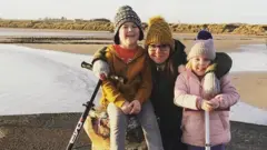 Esther Graham Esther Graham is in her thirties and is on a pier, crouching down with her arms around her two children who are about three or four years old. She is wearing a yellow hat, has glasses and a brown jacket. The boy, with a yellow jacket and grey bobble hat, is sitting on a concrete bollard. The girl, who is wearing a pink jacket and hat is standing beside it. Behind the trio there is a big beach with the sand dunes fringing it.