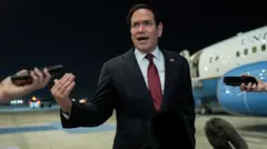 A man gestures with his right hand wearing a dark suit in front of an American government plane, microphones enter the frame.