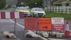 Two garda cars are parked on a road. A barrier is blocking the road and three signs are up. One says in Irish bóthar dúnta, and in English road closed. A cone and sandbags are on the road.