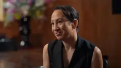 Gleit wears a sleeveless black blazer, and is sitting on a chair in front of a large glossy wood panelled room. A large vase of colourful flowers is also blurred in the background behind her.