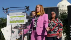 Reuters Liz Stein, who was a victim of late financier Jeffrey Epstein, speaks on the day of a rally in support of Epstein's victims, on Capitol Hill in Washington, DC, in September 2025. Liz is wearing a pink suir and standing in front of a podium with the word stand with survivors on a sign.