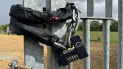 A close up of a large padded chain lock with a combination code wrapped around metal gates. Behind the gates an expansive green sports field can be seen.