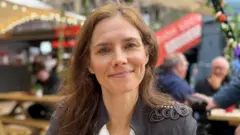 Amanda Knox. A 39-year-old woman with brown hair and wearing a black jacket and white shirt smiles at the camera. She is sitting in a pub garden surrounded by fairy lights and picnic benches.