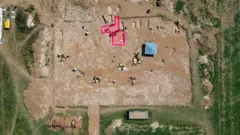 Kevin Church/BBC News Aerial view of the archaeological dig site. The location of a possible building has been graphically highlighted in pink. Six or seven graves are clustered around it. There are another six or seven graves visible that are located further from the highlighted feature. Archaeologists are working at different parts of the site wearing yellow high visibility jackets. The wider area of the cemetery that is being excavated has exposed brown soil. It is surrounded by a green field.