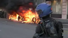 Police officer wearing protective gear watches car in flames