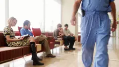 Stock photo of a young woman, a middel are man and a male and female couple with an infant  waiting in a hospital on chairs. A male doctor in blue scrubs walks along the corridor  right hand side. 