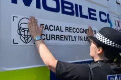 A police officer sticks a sign reading Facial Recognition Currently in Use on he side of a police van.