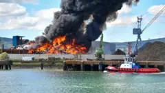 A large fire with a plume of black smoke rising from it is visible at Belfast Harbour. Part of the sea is visible in front of the fire, with a small boat docked to the right of the image. There is a blue sky, with some clouds, above. 