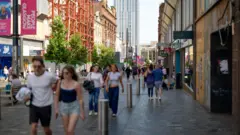 Pedestrians walk along a busy shopping street in Glasgow city centre, with retail stores, trees, construction scaffolding and modern high-rise buildings lining the pedestrianised thoroughfare.