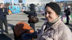 Elina wears a black bandage on the head, while her right hand is placed on the edge of the stroller. People in the background can see how to take pictures of each other